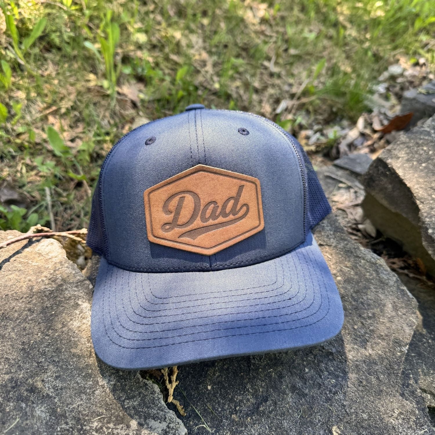 | BLACK-001-008, CHARC-001-008, NAVY-001-008, HGREY-001-008, MOSS-001-008, BROWN-001-008

Blue cap with 'Dad' leather patch on a stone surface with grass in the background