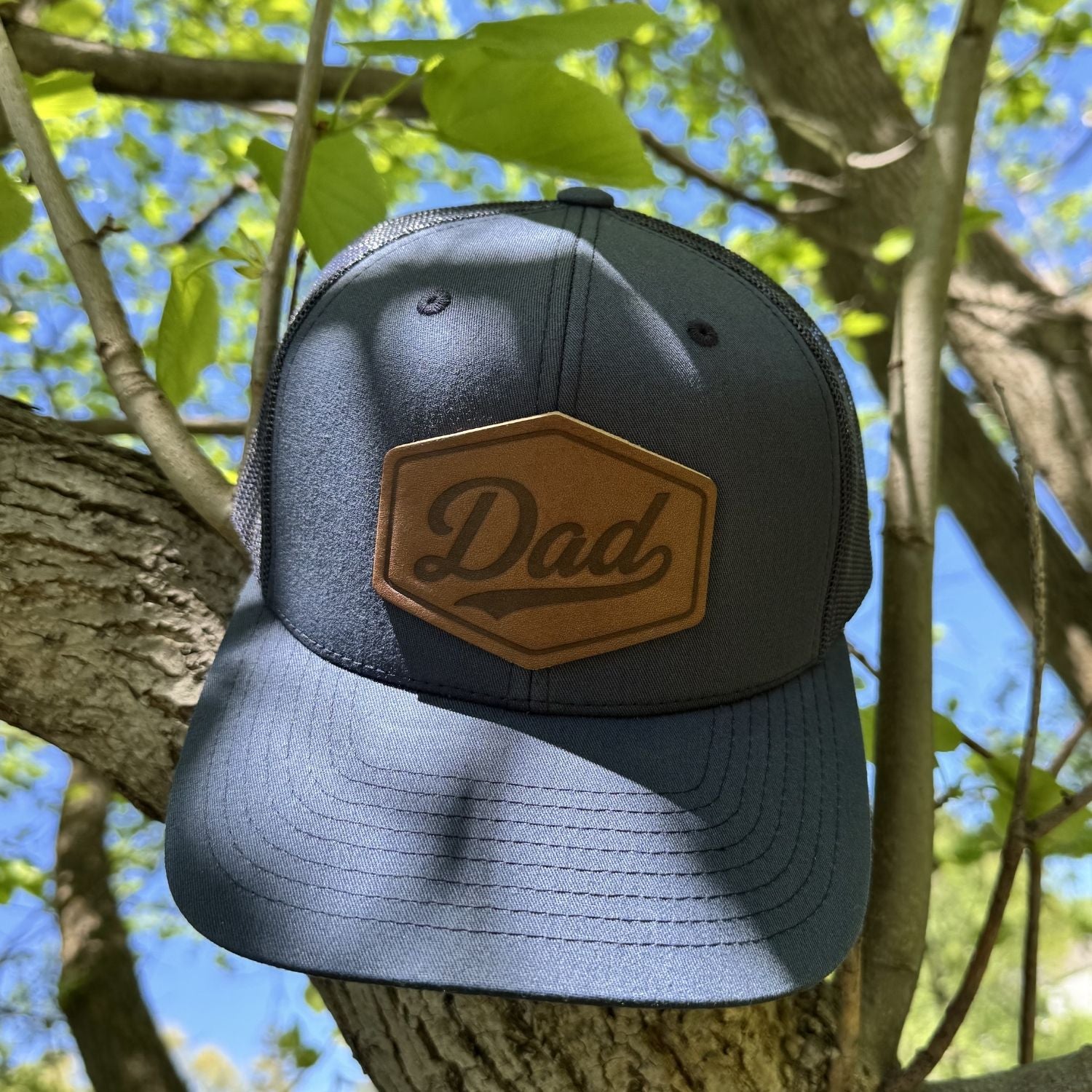 | BLACK-001-008, CHARC-001-008, NAVY-001-008, HGREY-001-008, MOSS-001-008, BROWN-001-008

Navy trucker hat with 'Dad' patch on a tree branch