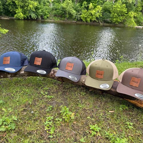 Five custom leather patch hats in various colors arranged on a log by a body of water.