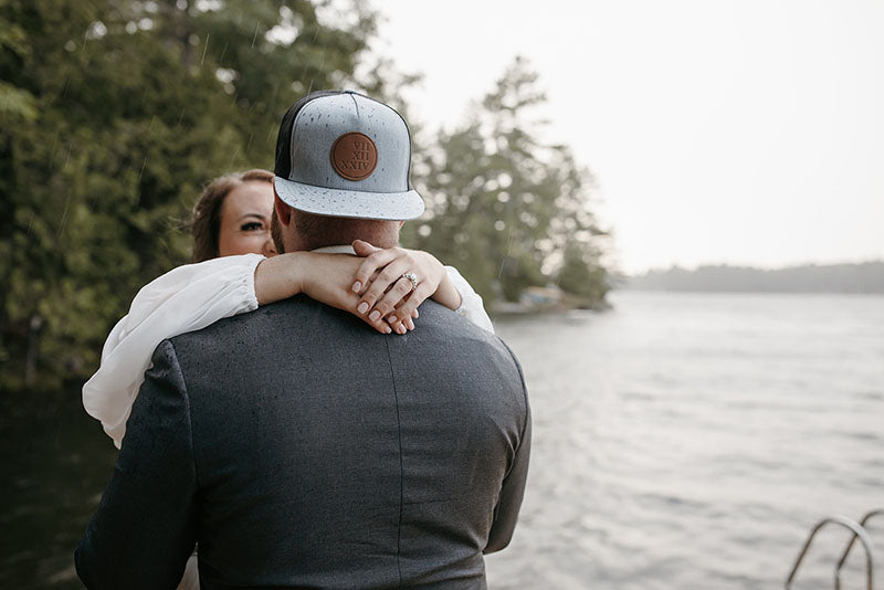 Two people embracing by a lake with trees in the background, wedding photo with a custom leather patch flat brim hat